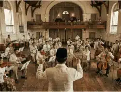 Latihan orkestra Indonesia modern dengan pengaruh musik klasik The Netherlands dalam gedung bergaya kolonial.